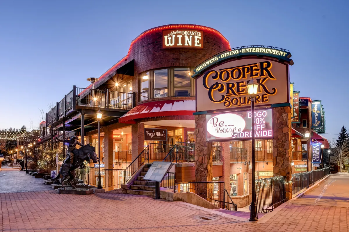 This image captures the vibrant Cooper Creek Square, featuring visible signage, a wine shop, stairs leading to an upper deck, and a horse sculpture. People can be seen enjoying shopping, dining, and lively entertainment in a festive, urban mountain setting.