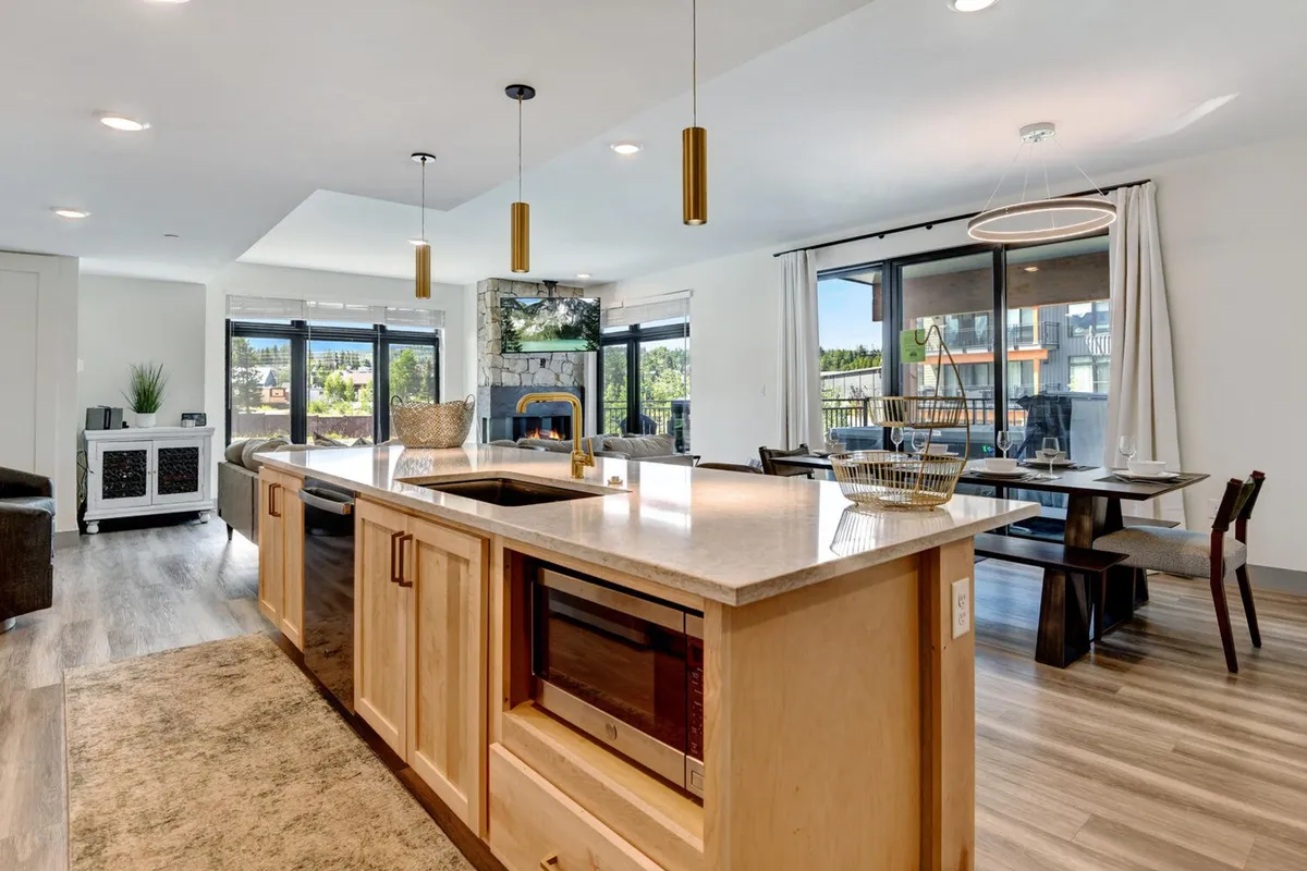 A contemporary open-plan space featuring a kitchen island, dining table, fireplace, and large windows. The room is bathed in natural light, inviting both relaxation and lively conversation in a chic mountain setting.