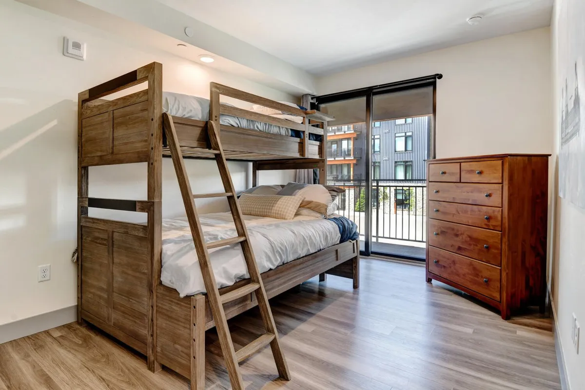A playful bunk room with a twin-over-queen bunk bed, matching bedding, a dresser, and a large window streaming in morning light. The room feels cozy and inviting, perfect for children or groups. The arrangement encourages socializing and shared experiences, while the natural light ensures a cheerful atmosphere.