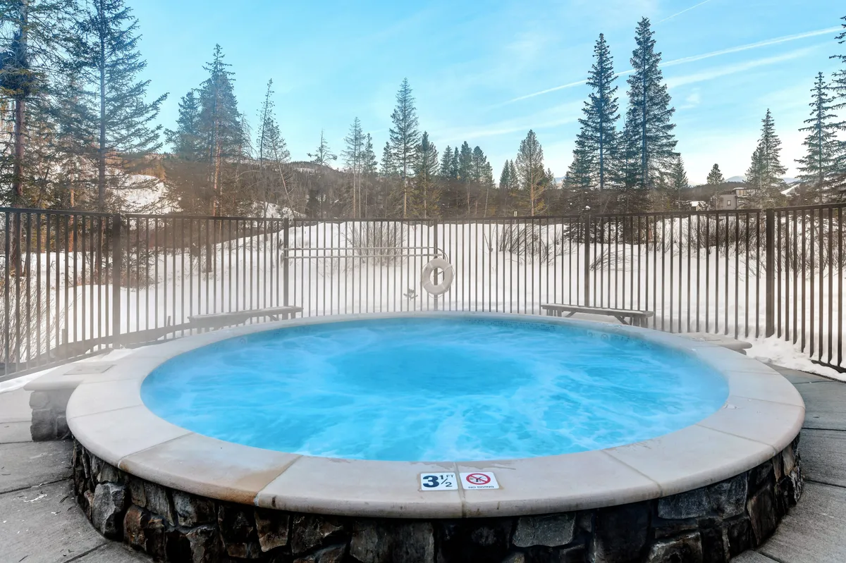 This exterior image showcases a private hot tub nestled amidst snowy surroundings and pine trees. People are seen relaxing in the hot tub, taking in peaceful nature views, suggesting a serene retreat under the open sky.