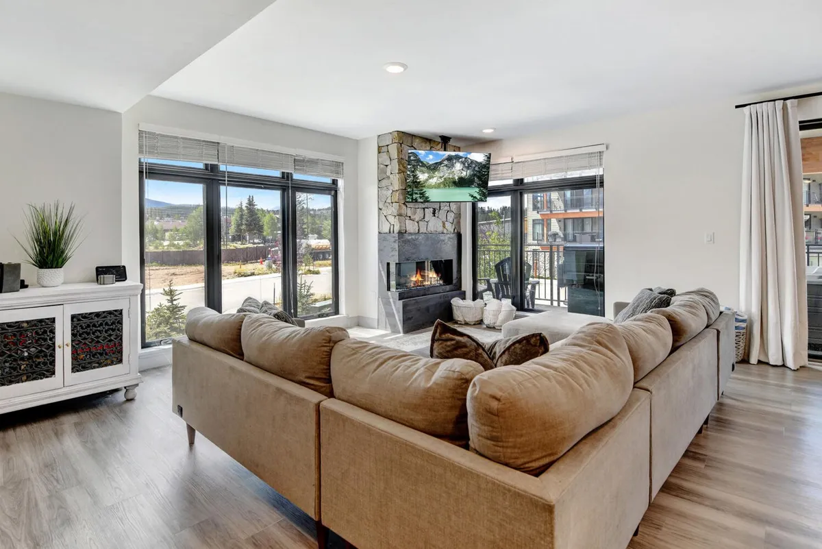 This inviting interior shot features a plush sectional sofa positioned by a modern, color-changing fireplace, with a large window framing scenic mountain views. The wall-mounted TV and stylish décor create a cozy, contemporary gathering space.