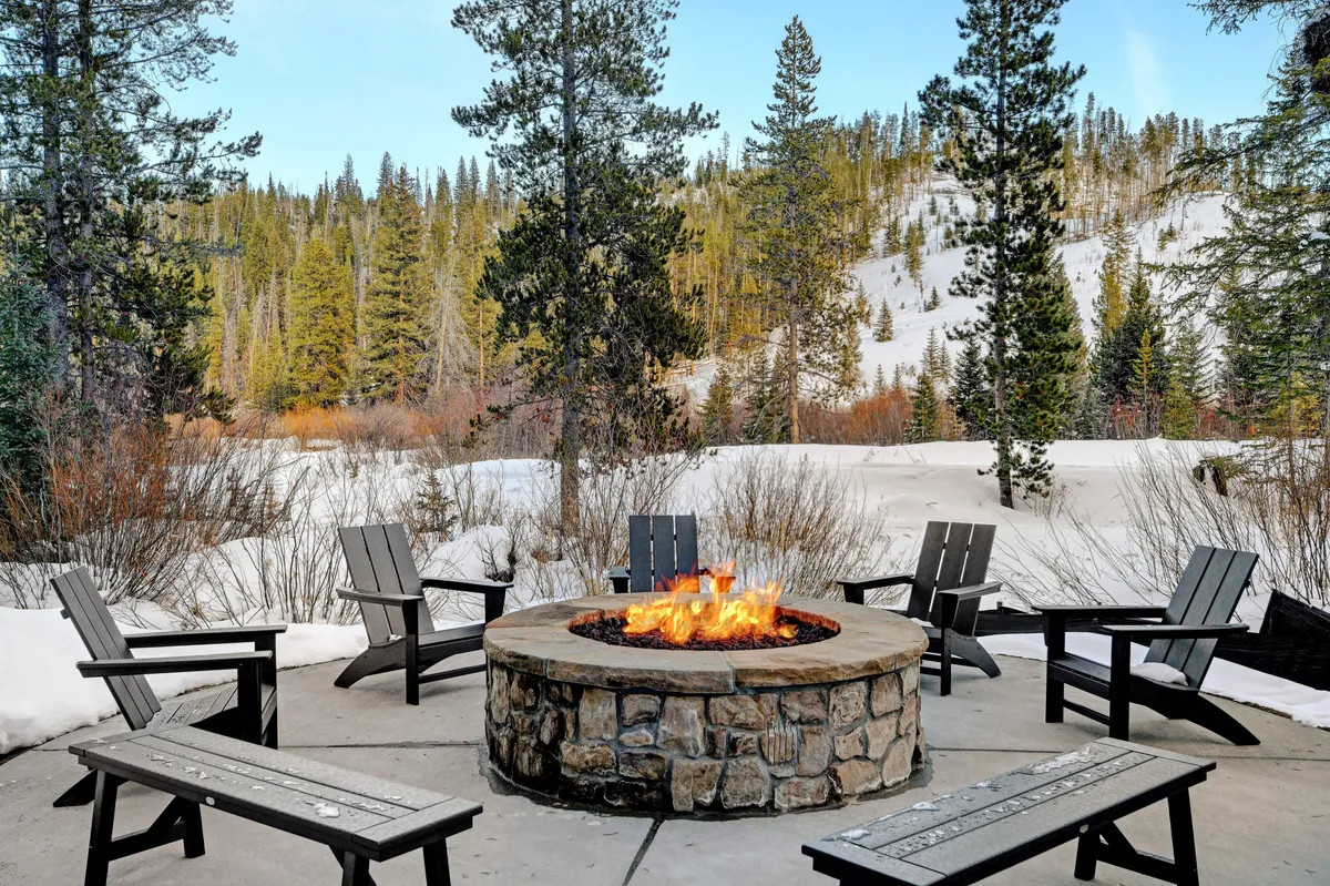 An exterior scene featuring a glowing fire pit with surrounding seating set in a snow-covered landscape with tall pine trees. People gather around, enjoying warm drinks and sharing stories under the stars.