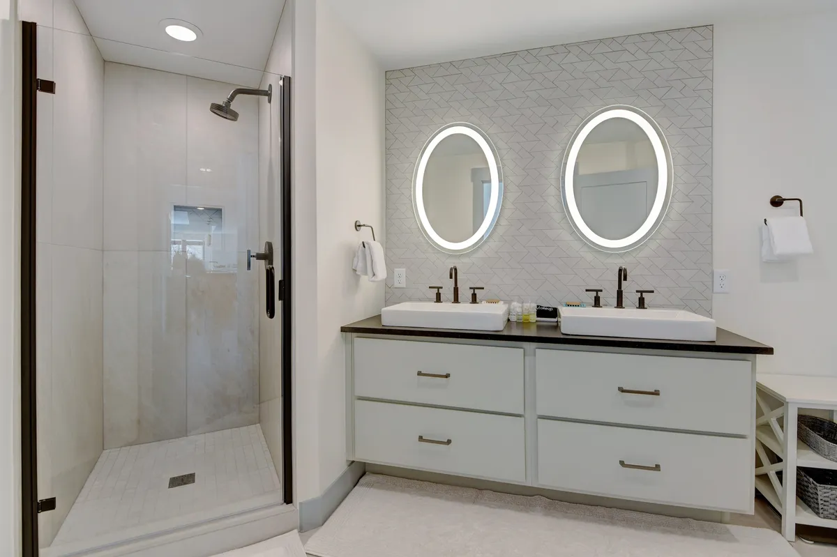 A modern bathroom with double Jack & Jill sinks, wide mirrors, and a glass-walled shower area. The room is bright, with clean lines and spa-like finishes, evoking a sense of privacy and relaxation for guests.