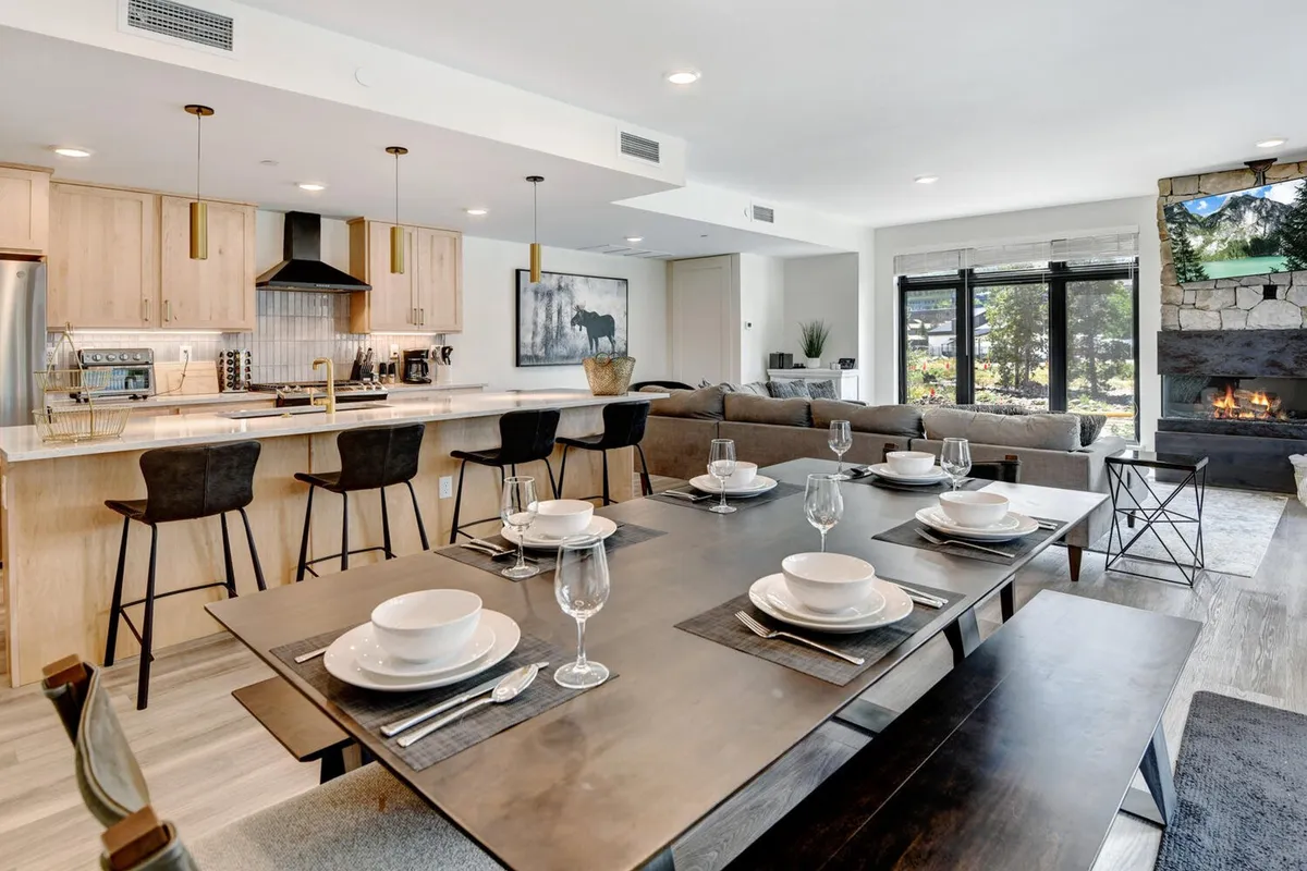 A beautifully arranged dining area with a large table set for a meal sits adjacent to a kitchen island with bar stools, all anchored by a striking stone fireplace. The open-concept layout encourages group gatherings and communal dining.