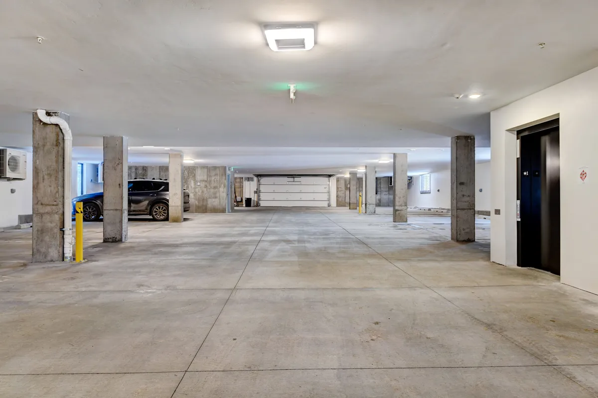 An interior view of a secure, well-lit parking garage with concrete pillars, parked cars, and an elevator. The scene is practical and emphasizes convenience and safety for guests’ vehicles.