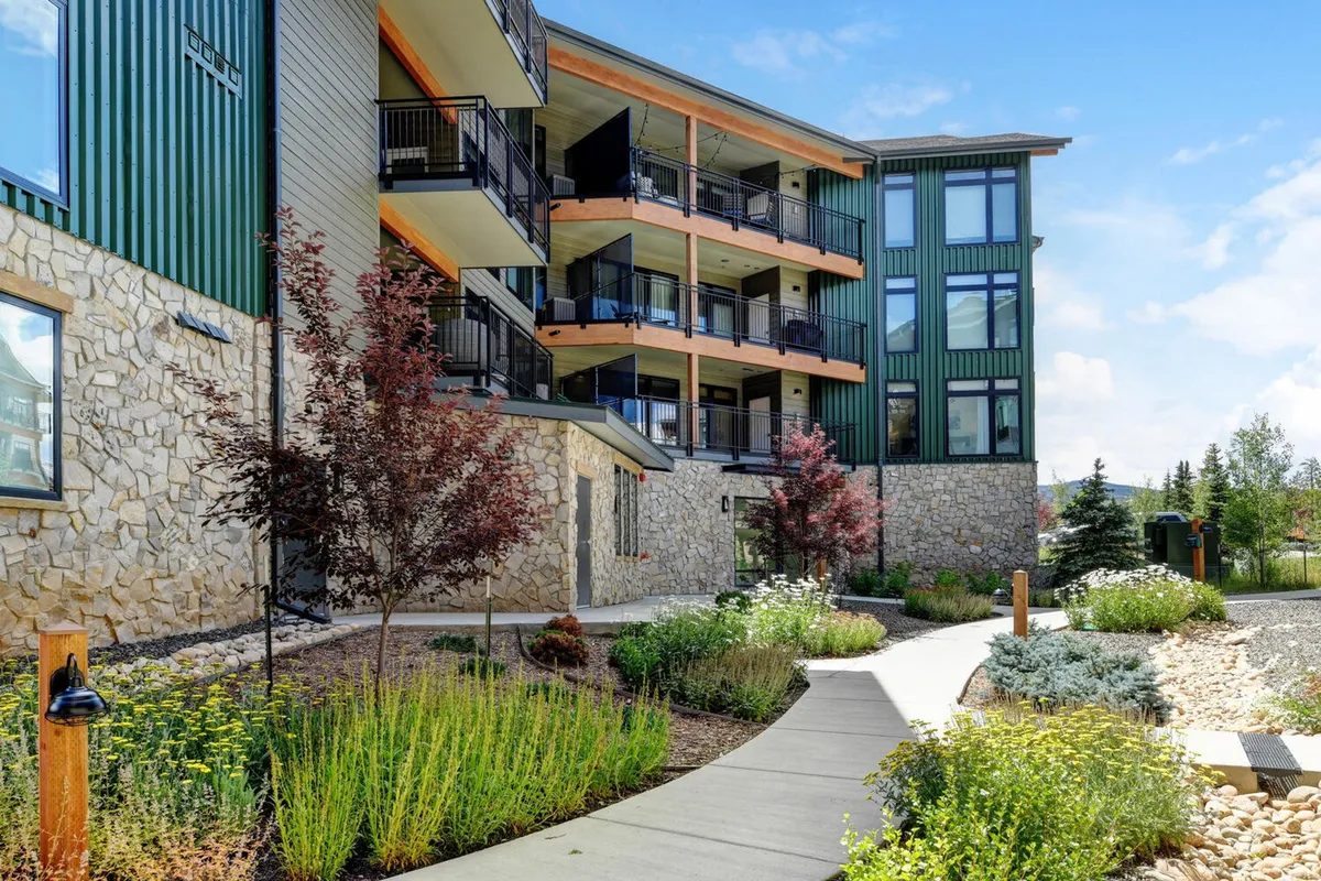 An exterior view of the property shows stone walls, green siding, balconies, and landscaped gardens, with pathways inviting guests towards the entrance. The scene conveys a sense of tranquility and well-maintained surroundings.