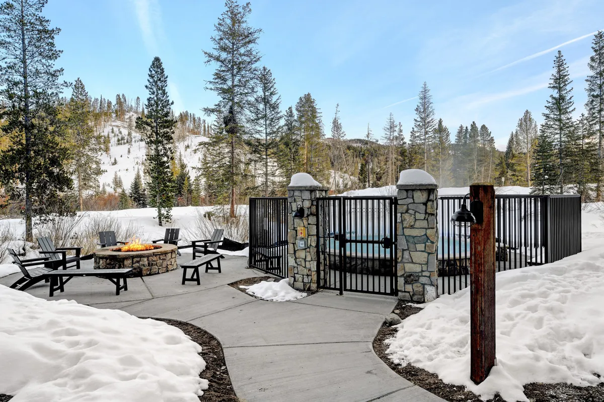 A welcoming outdoor scene with a fire pit surrounded by chairs, a bubbling hot tub, snow-covered ground, and towering trees. The setting glows with warmth and relaxation, perfect for soaking or gathering after a day of adventure.